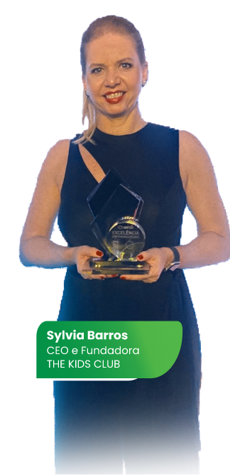 Mulher feliz, de vestido preto sem mangas, segurando um troféu em suas mãos. No final da imagem, tem um box verde com os cantos arredondados escrito Sylvia Barros, CEO e Fundadora THE KIDS CLUB